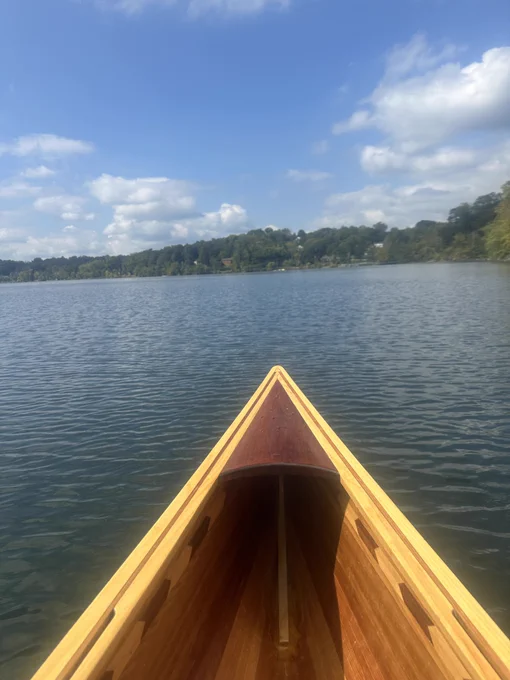 The bow of a cedar strip canoe is center frame. The photographer must be sitting in the bow. Ahead are calm waters of a reservoir lined by green trees all around.