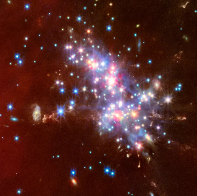 At centre is a compact star cluster composed of luminous red, blue, and white points of light. Faint jets with clumpy, diffuse material extend in various directions from the bright cluster. Translucent red wisps of material stretch across the scene.