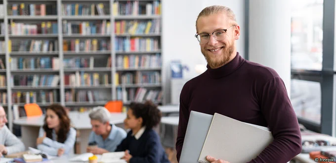 Bild zeigt eine männl. gelesene Person im Vordergrund, mit Bart, Brille und dunklem Rollkragenpullover, einen Laptop und Papierblock haltend. Im Hintergrund sieht man unscharf vier sitzende Personen und viele Bücher.
Bild von FreePix