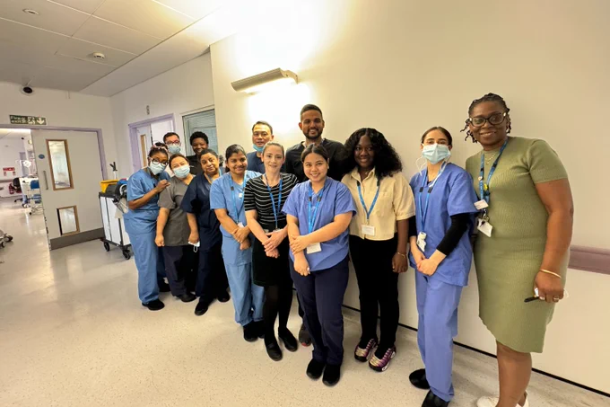Team photo of the clinical haematology team at Hammersmith Hospital. Members of staff are standing in a hospital corridor and smiling towards the camera.