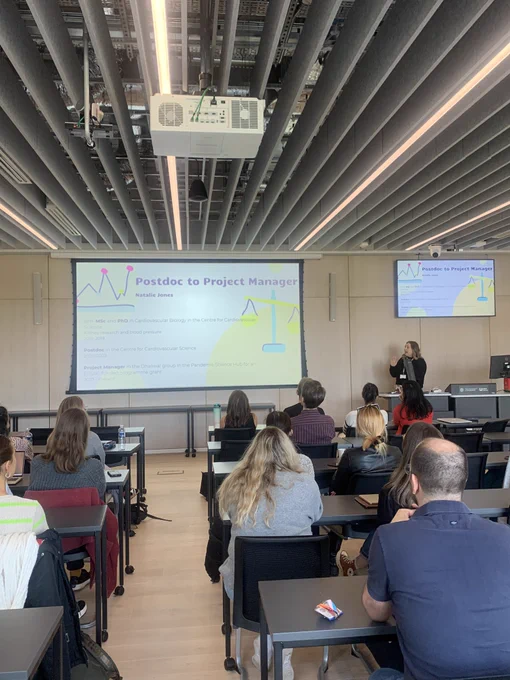 Natalie presenting in front of her slides with the title “Postdoc to Project Manager” in a room full of people.