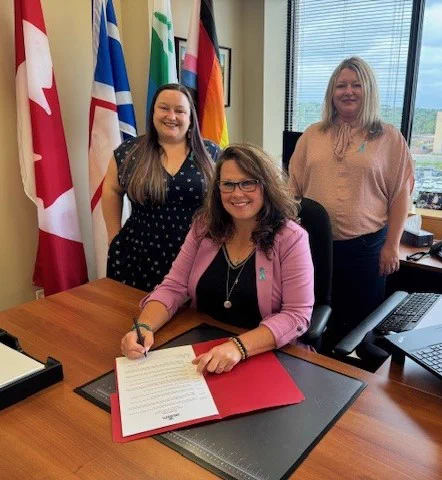 Women and Gender Equality Minister Pam Parsons sitting at her desk signing the proclamation declaring September 15-21 as Sexual Violence Awareness Week in Newfoundland and Labrador.  Also pictured are Mary Walsh, Executive Director of End Sexual Violence NL on the left and Dawn Nolan, Operations Coordinator with End Sexual Violence NL on the right.