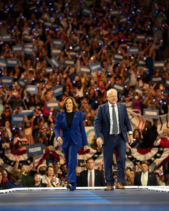 Vice President Kamala Harris and Governor Tim Walz waving to a crowd on a stage in a packed arena. 
