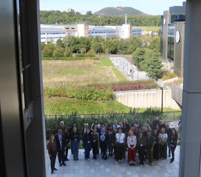 A photo of a large group of people in front of the Usher Institute building in the Bioquarter. You can see IRR South and the Royal Infirmary Hospital behind them, and Craigmillar Castle and Arthur’s seat in the background, under a blue, cloudless sky.