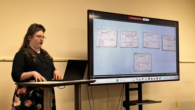 A person giving a presentation beside a laptop, with a large screen displaying WWII Ration Books behind them.