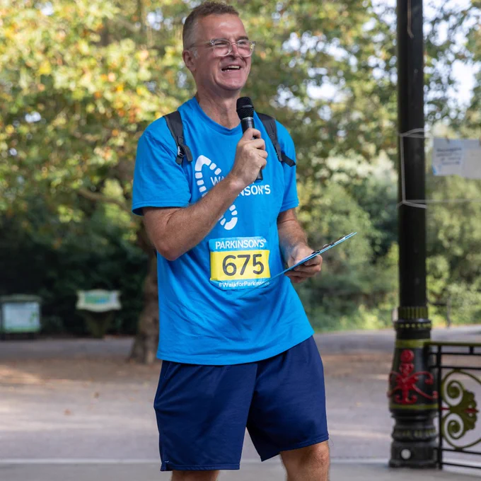 Dave Clark in a blue Parkinson's UK t-shirt speaking into a microphone and holding a clipboard in a park setting.