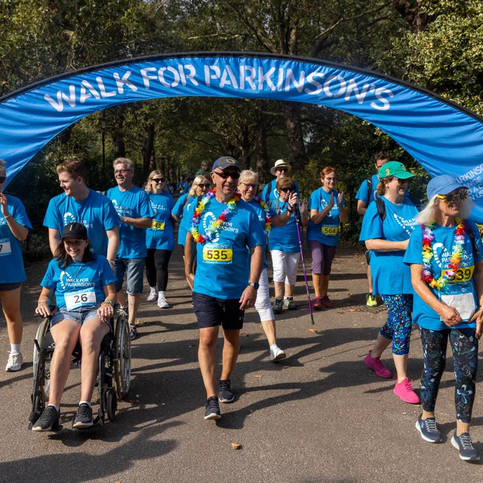 Group of participants walking under a blue 'Walk for Parkinson's' arch at a charity event, including one person in a wheelchair, all wearing event t-shirts and bib numbers.