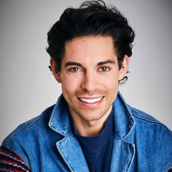 Portrait of a Tom Read Wilson with dark hair wearing a denim jacket and a colorful sweater.