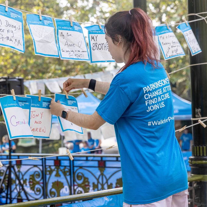 Person in a blue "Parkinson's Change Attitudes Find a Cure Join Us" t-shirt pins notes on a line at a public event.
