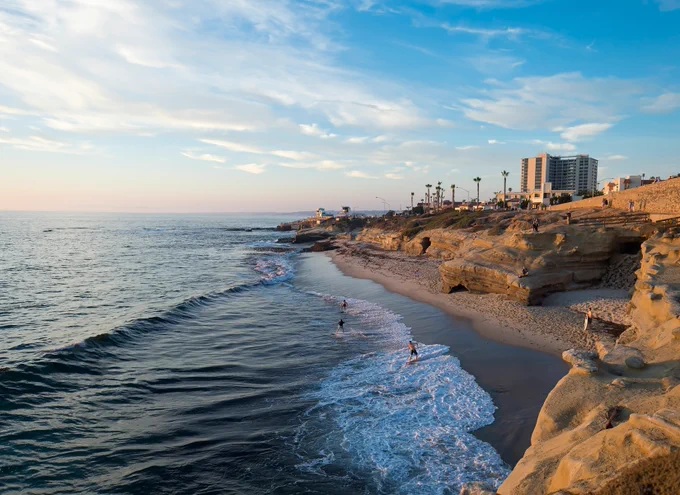 A photo of the California coast with waves crashing into the beach and people walking on the beach. The sky is blue and partly cloudy. A tall building is in the background.