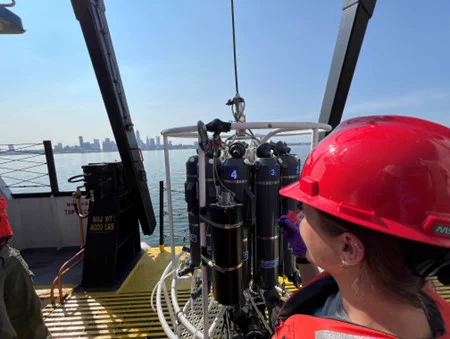 The crew aboard the R/V Lake Guardian deploy the first rosette of the CSMI survey. Crew members stand in red safety hats and orange life jackets on the deck of the Lake Guardian, at the large rosette tool. The Cleveland Skyline is in the background. 
