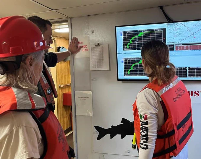 Crew members aboard the R/V Lake Guardian assess a screen with graphs displayed on it. They wear orange life jackets. 