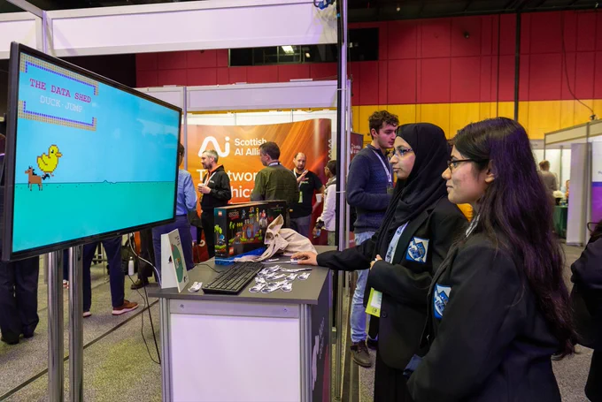 two woman engaging with a digital game at an exhibitor stand. One woman is playing with the computer and the woman beside her is watching