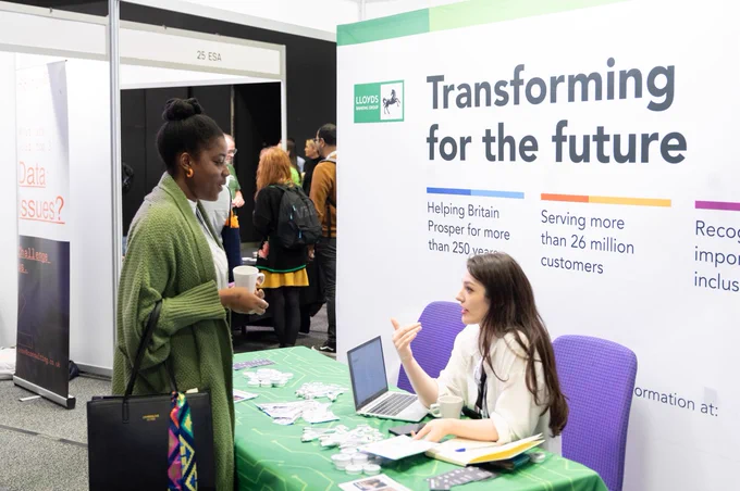 woman sat down behind LLoyds exhibitor stand talking to woman who is standing on the other side