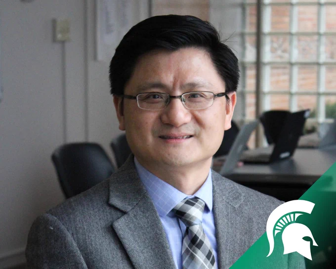 A professional headshot of a man wearing glasses, a gray suit jacket, a light blue dress shirt, and a patterned tie. He is smiling slightly while sitting in an office setting with large windows in the background. The Michigan State University Spartan logo is visible in the bottom right corner of the image.