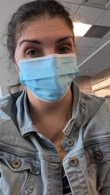 Latina woman with her brown hair pulled into a bun smiles at the camera but you can't really tell because she's wearing a blue surgical mask. She has on a light wash denim jacket.