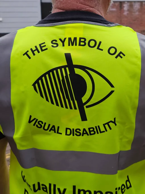 A close up photo of the back of a man wearing a high visibility vest for visually impaired people. The symbol of visual disability is at the centre of the man's back. 