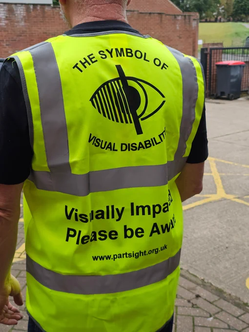 A photo of the back of a man in a high visibility vest for visually impaired people. The symbol of visual disability is on the centre of his back, and the words "visually impaired please be aware" are underneath
