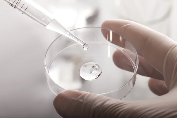 The image shows a close-up of a laboratory setting, where a hand wearing a latex glove is holding a clear petri dish. A pipette is positioned above the dish, and a droplet of liquid is about to be released into the dish, where another droplet is already present. The background is blurred, emphasising the precision and cleanliness of the scientific process.