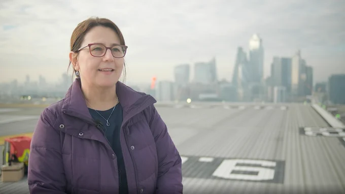 A woman stands on a helipad located on the roof of a hospital. Over her shoulder is the city of London.