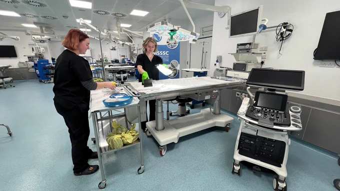 Two females in black scrubs prepping a surgical theatre.