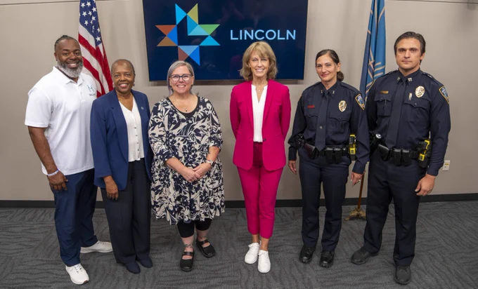 Group of six individuals posing for a photo at a Lincoln event, including two police officers in uniform and four civilians. American flag and Lincoln logo visible in the background.