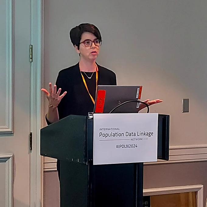 Dr. Jennifer Brooks, a woman with short dark hair and glasses, speaks at a podium at the IPDLN Conference. 