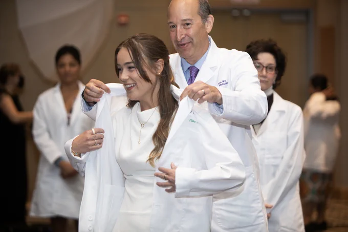 A Rush Medical Student smiles while being assisted in putting on a white lab coat by another individual, at the white coat ceremony.