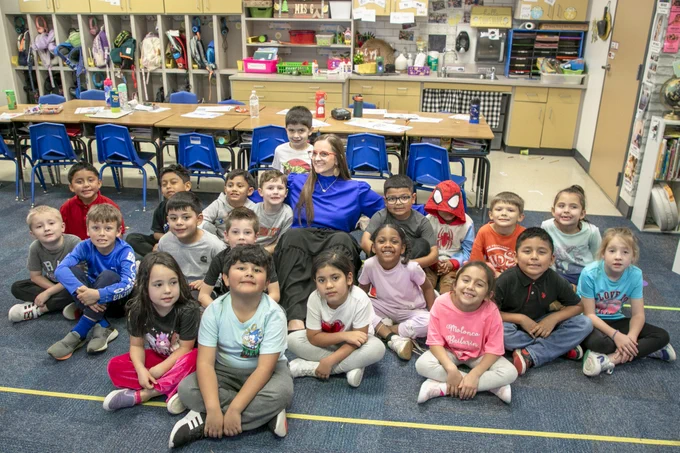 A teacher sitting on the floor with a group of her students, smiling at the camera. 