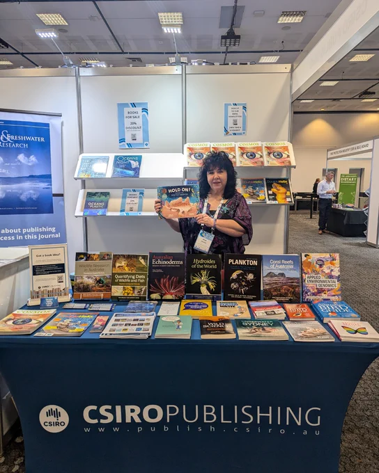 Gina Newton standing at the CSIRO Publishing conference booth behind a table full of books, smiling at the camera and holding her book Hold On! Saving the Spotted Handfish.