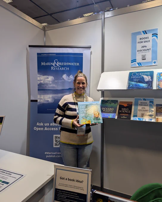 Prue Francis standing at a conference booth, smiling at the camera and holding her book The Great Southern Reef.