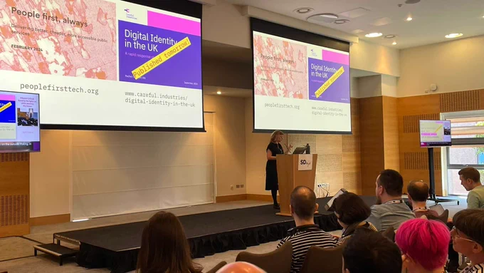 A speaker at a podium presenting on "Digital Identity in the UK" at a conference, with the audience listening attentively. The presentation slides are visible on screens showing graphics and text related to digital identity.