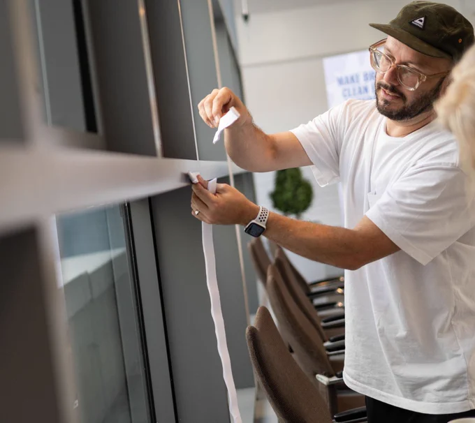 Person in a white shirt and cap adjusting a strip of paper on a glass wall at SDinGov