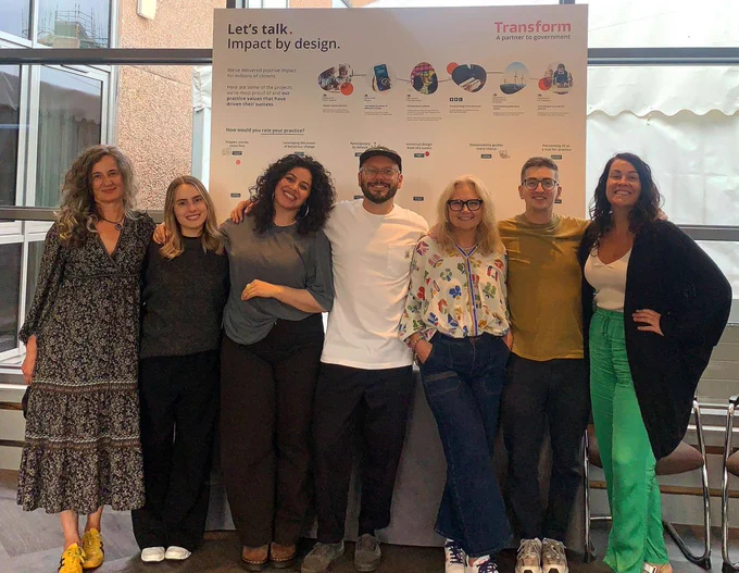 Group of seven individuals standing and smiling in front of a poster titled "Transform. Impact by design.