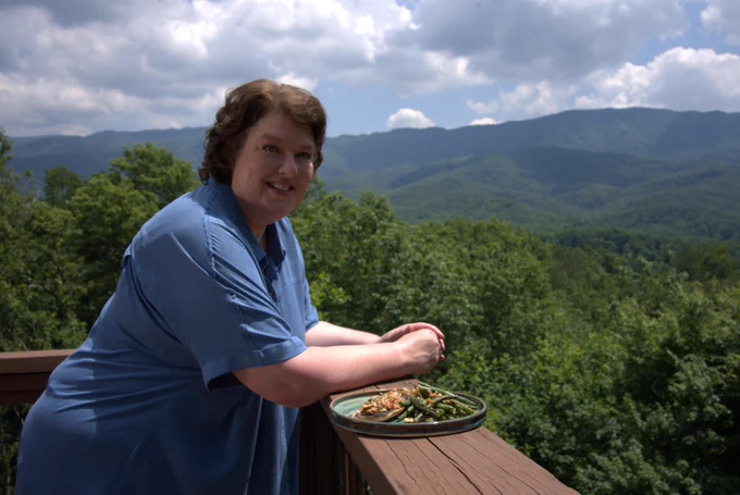 A smiling woman with short brown hair wearing a blue short sleeved shirt leans over a wooden balcony with a plate of food beside her and a wooded landscape in the background 
