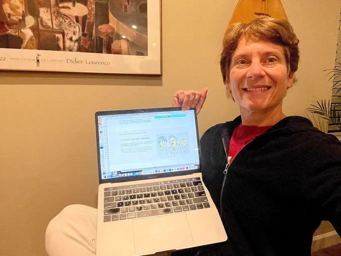 A selfie by Carolyn Bertozzi at home. She is holding up a laptop with the homepage to NobelPrize.org showing the 2022 chemistry prize announcement.