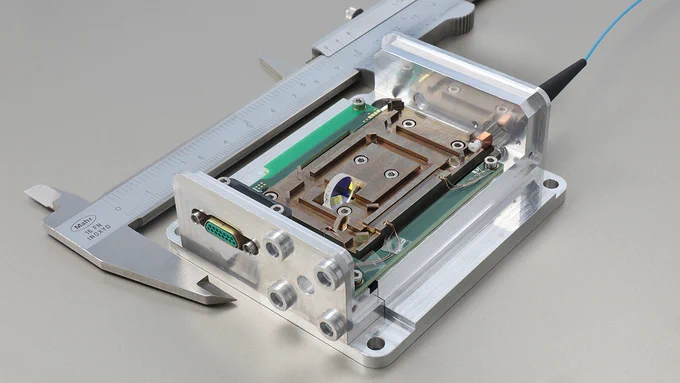 The photo shows a laser module next to a measuring caliper, which illustrates that the module is no larger than 10 cm. Various lenses and a crystal can be seen in the module. The module is coupled with a fiber that guides the pump radiation into the module.