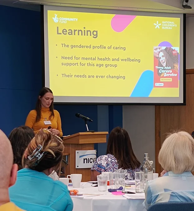 Image shows Andrea, wearing a mustard coloured rollneck, standing behind a lectern addressing the room. A powerpoint slide, entitled 'Learning', is behind her.