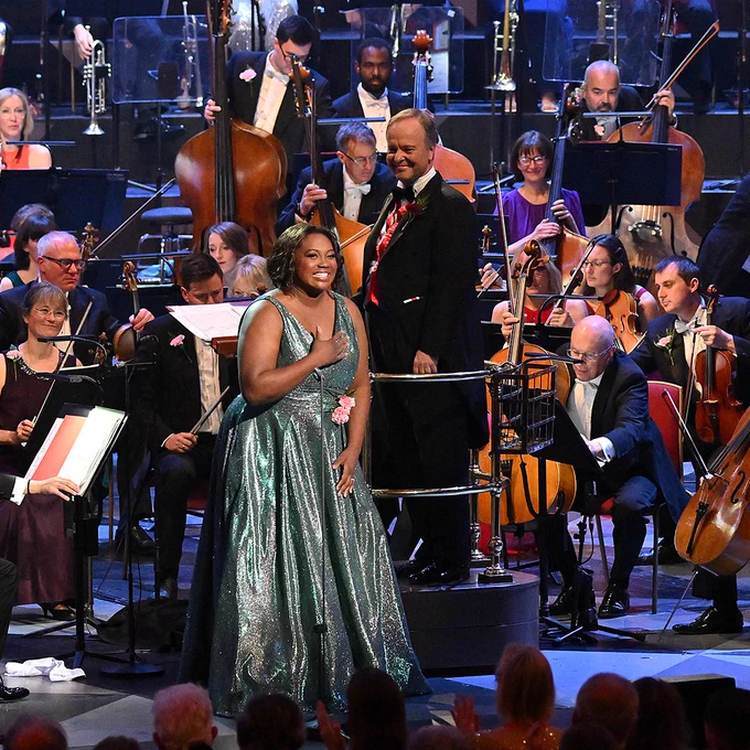 Photo of singer stood in the middle of a large orchestra. The conductor and musicians of the orchestra look to the singer, as the singer smiles to the audience. 