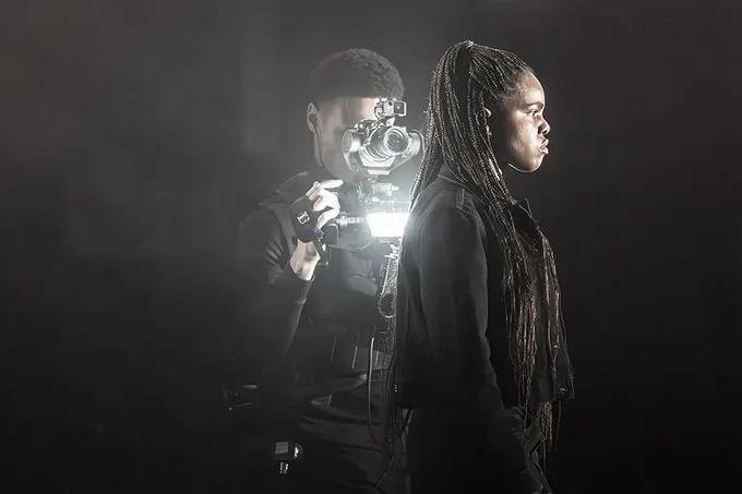 A photo showing a performer on a darkened stage, lit by a camera operator obscured by shadow 