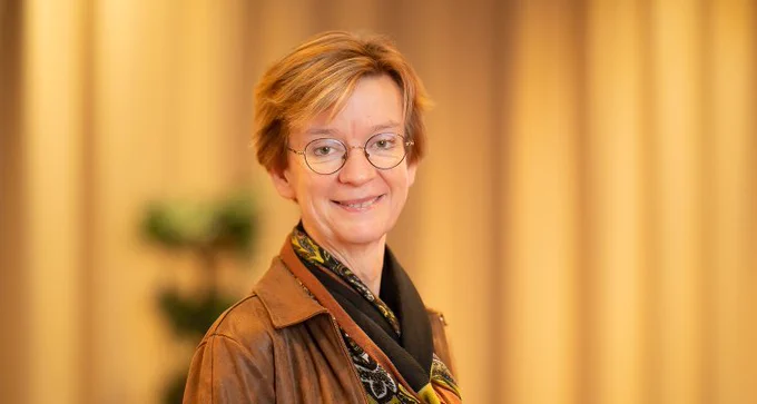 "A smiling woman with short, light brown hair, wearing glasses and a brown jacket over a patterned scarf. The background is softly blurred with warm, beige tones."