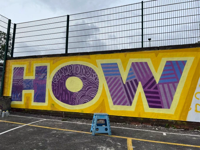 Part of a mural at a basketball court with the word HOW in purple against a yellow-mustard background.
