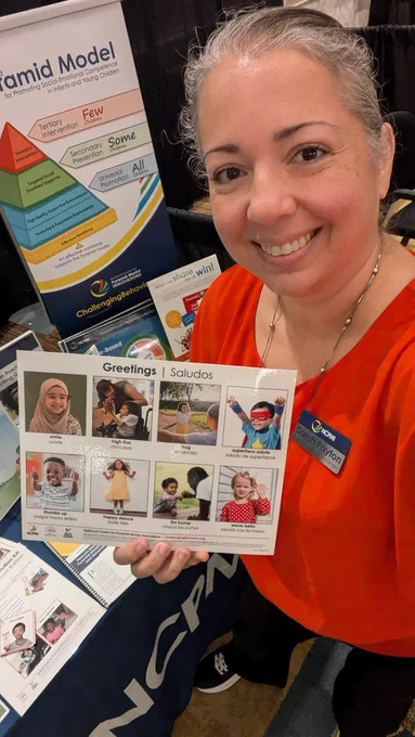 Sarah at the Division for Early Childhood (DEC) conference holding a Greetings/Saludos card from a resource kit, standing in front of the Pyramid Model sign by the NCPMI table.