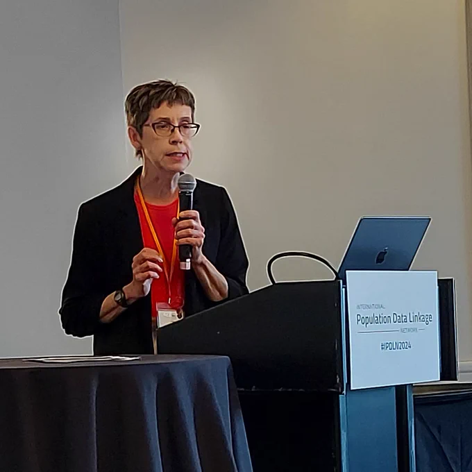 Lisa Lix, a woman with short hair and glasses, holds a microphone while speaking at a conference podium.