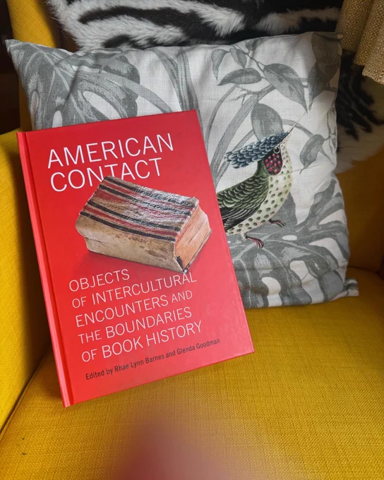 Red hardcover in sans serif font AMERICAN CONTACT: OBJECTS OF INTERCULTURAL ENCOUNTERS AND THE BOUNDARIES OF BOOK HISTORY edited by Rhae Lynn Barnes and Glenda Goodman (@pennpress). It leans against a cushion with a bright tropical bird on a mustard yellow chair