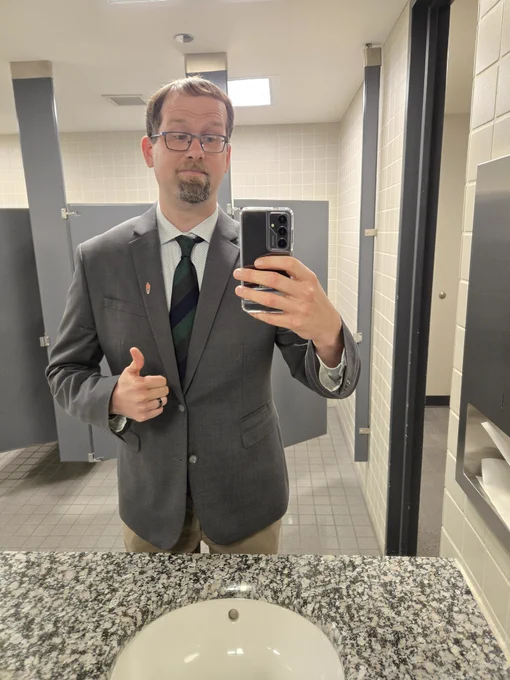 Man in suit taking picture of himself in bathroom mirror giving thumbs up