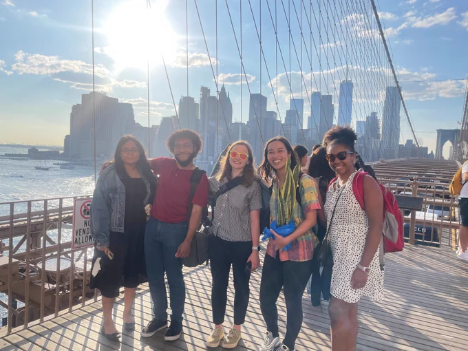From left to right: BetaNYC's Naeema, Dimitri, Kate, Jazzy, and Gabby pose together on the Brooklyn Bridge. The Manhattan skyline and sunshine illuminate the background.