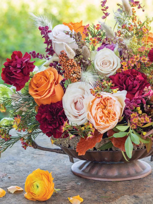 An arrangement in a pedestal basin including garden roses, carnations, ranunculus, waxflowers, and bunny tail grass in tones of orange, burgundy, peach, and yellow. 