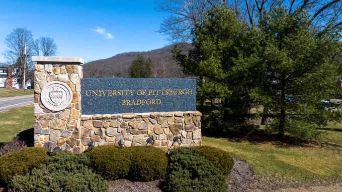 Entrance sign of University of Pittsburgh Bradford, featuring a stone structure with the university seal, under a clear sky.