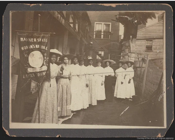 A sepia photograph of 9 African-American women posed, standing with Nannie Burroughs, holding a banner that reads, [Banner State Woman's National Baptist Convention].